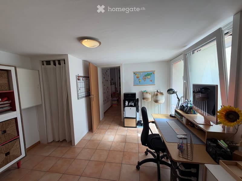 A well-lit, furnished room with a desk setup, a chair, a keyboard, a mouse, a lamp, a shelf, a printer, a radiator, a curtain, a world map, a vase with a sunflower, and a shelf with various items.