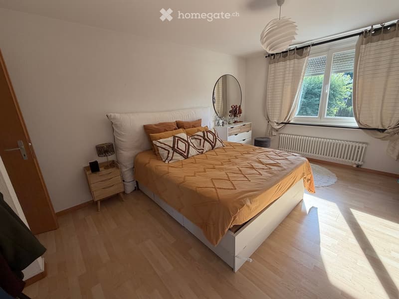 Bright bedroom with wooden flooring, large bed with brown bedding, nightstand with lamp, mirror, and window with curtains.