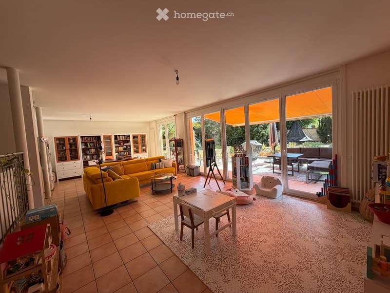 Bright living room with large glass doors leading to a patio, yellow sofa, coffee table, small dining table with chairs, bookshelf, and various toys.