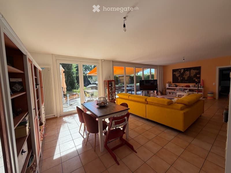 Bright living room with large glass doors leading to a patio, yellow sofa, wooden dining table with chairs, bookshelf, and television.