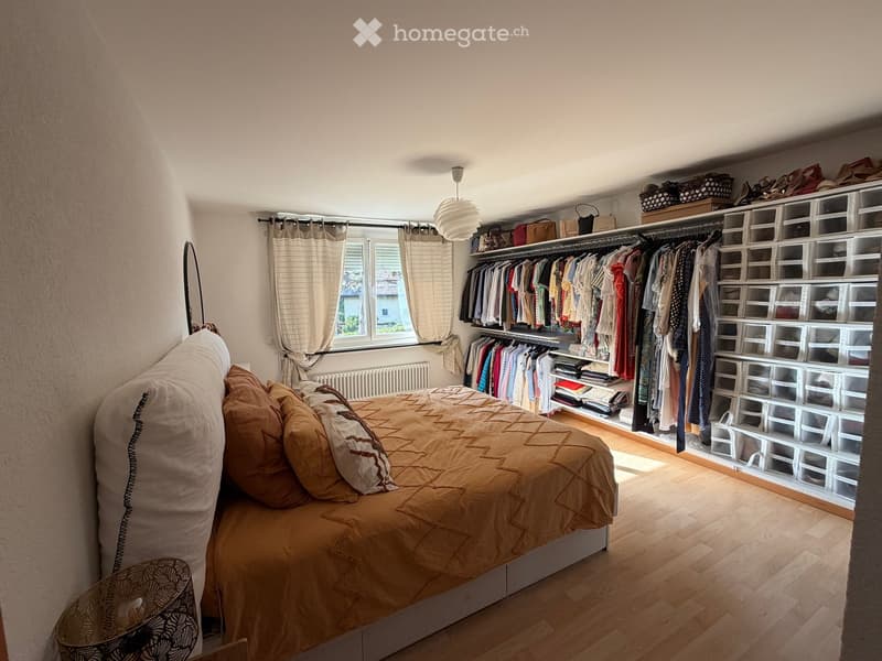 Cozy bedroom with a large bed, pillows, and a wardrobe with hanging clothes and storage boxes. The room features wooden flooring, a window with curtains, and a modern lamp.