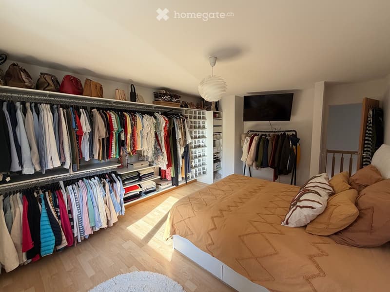 Well-lit bedroom with a large bed, a built-in wardrobe, and a clothing rack. The room features a pendant light, a screen mounted on the wall, and several handbags and baskets on the wardrobe shelves.