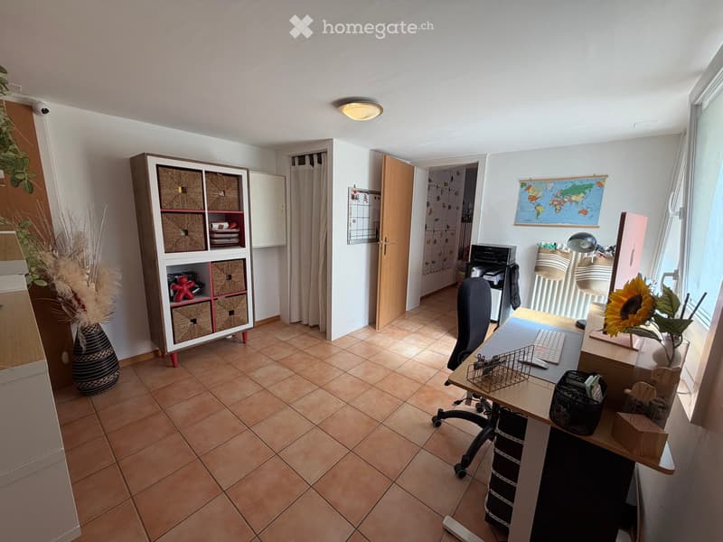 A well-lit, furnished room with a desk, chair, keyboard, screen, lamp, vase, shelf, printer, map, and curtain.