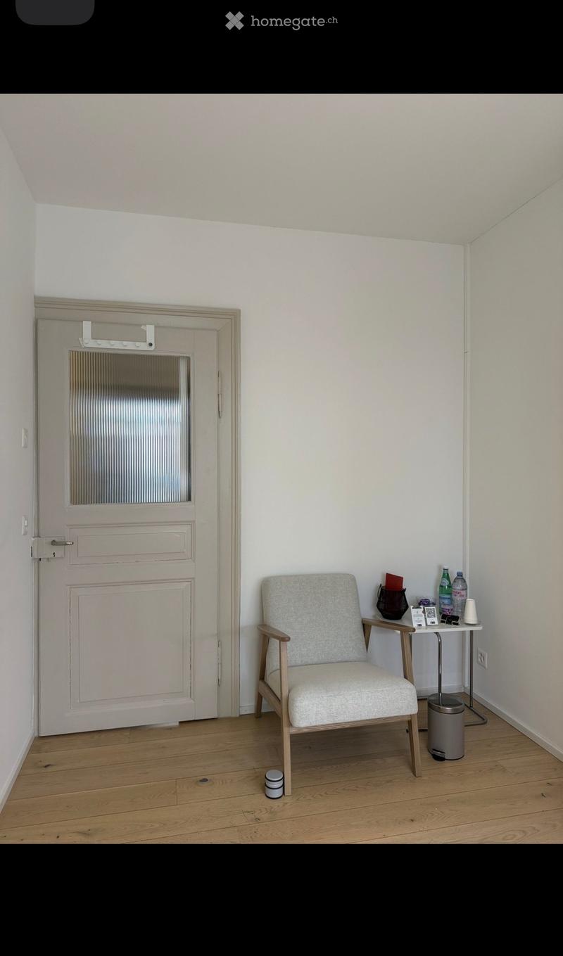 A small, furnished room with light wooden flooring, a single armchair, a small table with various items, and a door with a frosted glass panel.