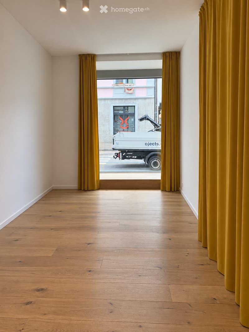 Empty room with wooden flooring, white walls, and large windows with yellow curtains, showing a view of the street outside.