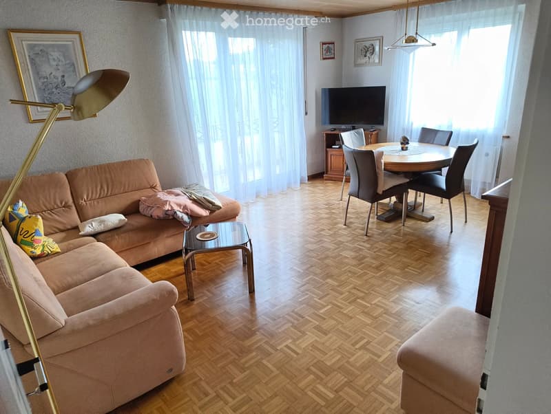 A well-lit living room with a beige sofa, coffee table, dining area with a round table and chairs, and a television on a stand. The room features wooden flooring and large windows with white curtains.