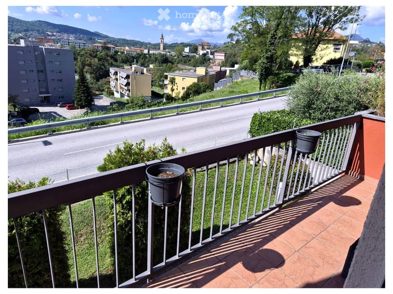 Balcony with metal railing, tiled floor, and two black planters, overlooking a street and surrounding buildings.