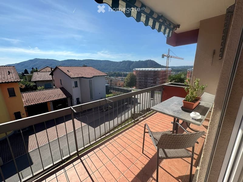 Balcony with a table, chair, and potted plant, overlooking a town with houses and mountains in the background.