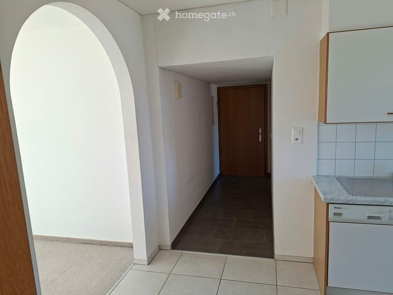 A narrow hallway with tiled floors, white walls, and a wooden door at the end. There is a light switch on the wall and a small cabinet with a countertop and a dishwasher visible on the right side.