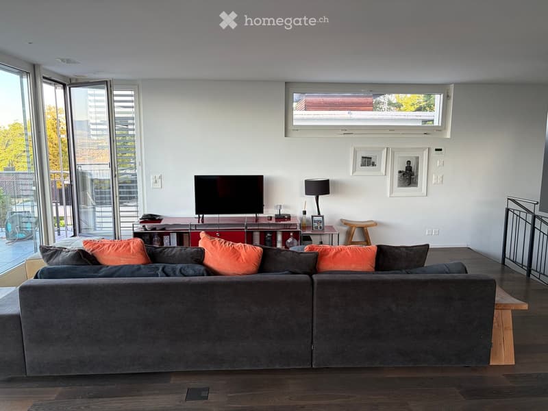 Modern living room with a large sectional sofa, orange and grey pillows, a wooden floor, large windows, and a television on a red stand.