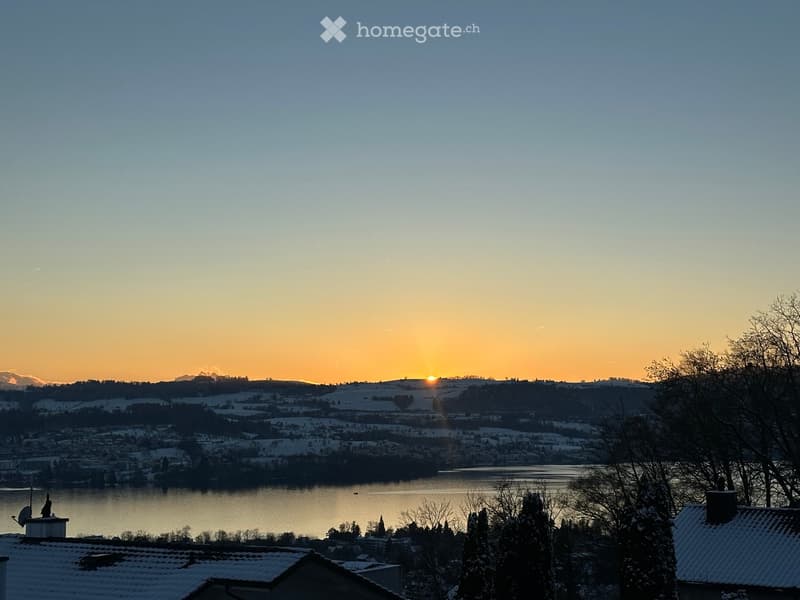Panoramic view of a snow-covered landscape at sunset, featuring a lake, distant mountains, and rooftops in the foreground.