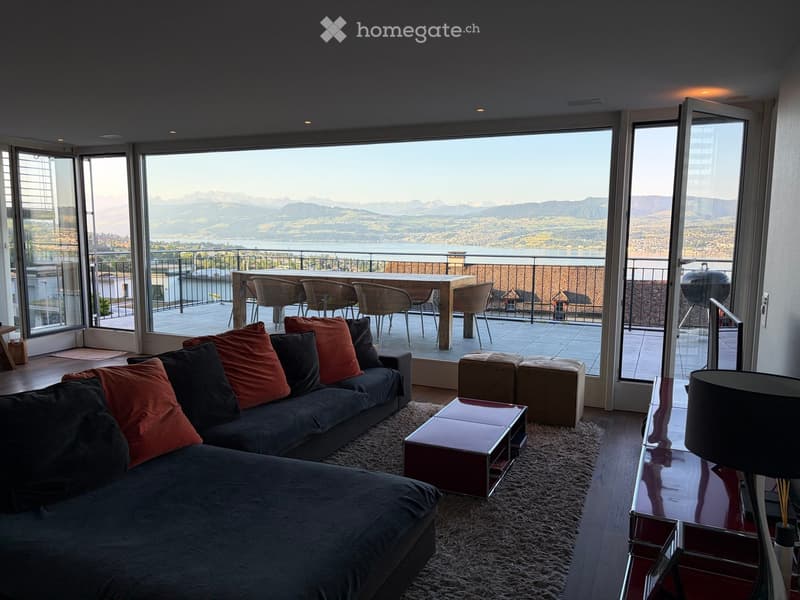 Modern living room with large windows offering a scenic mountain view, featuring a dark grey sectional sofa with colorful pillows, a sleek coffee table, and a small outdoor dining area with chairs and a table.