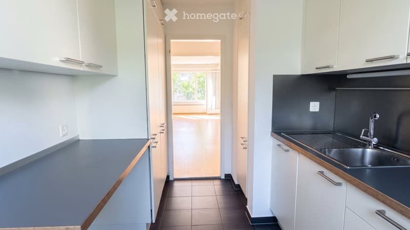 Modern kitchen with white cabinets, dark countertops, and a stainless steel sink. The space is clean and well-lit, with a view into an adjacent living area.