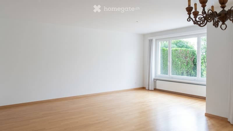 Empty room with wooden flooring, white walls, large window with white frame and curtain, radiator below window, and a chandelier hanging from the ceiling.