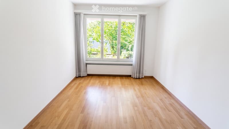 Empty room with light wooden flooring, white walls, large window with grey curtains, and a radiator below the window.