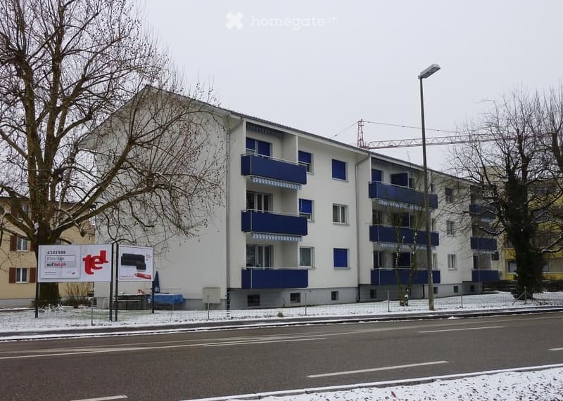 Multi-story residential building with blue balconies, white facade, and snow-covered surroundings.