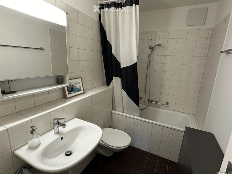 Modern bathroom with white tiles, a sink, toilet, bathtub, shower, and a mirror cabinet. Features a black and white shower curtain and a framed picture on the shelf.