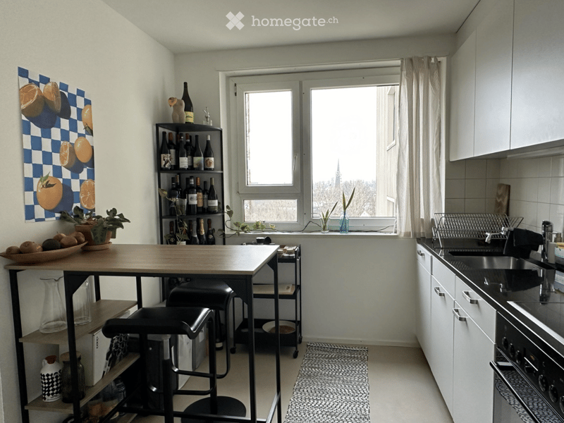Modern kitchen with white cabinets, a wooden table, two black stools, a sink with a faucet, an oven, a bottle rack, a bowl of fruit, a vase with a plant, and a painting on the wall.