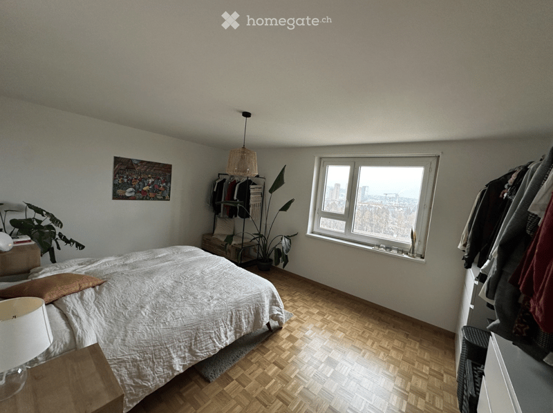 Cozy bedroom with wooden herringbone flooring, large window with city view, bed with white bedding, nightstand with lamp, wardrobe, and potted plants.