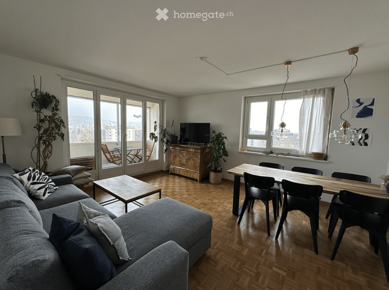 Modern living and dining area with wooden flooring, large windows, a grey sectional sofa, coffee table, dining table with chairs, TV, and potted plants.