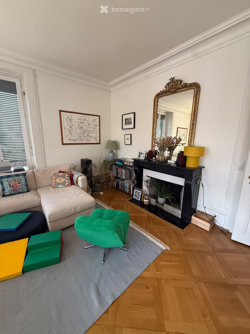 Living room with a fireplace, a large mirror, a sofa, an ottoman, a green chair, and decorative items on the mantel.