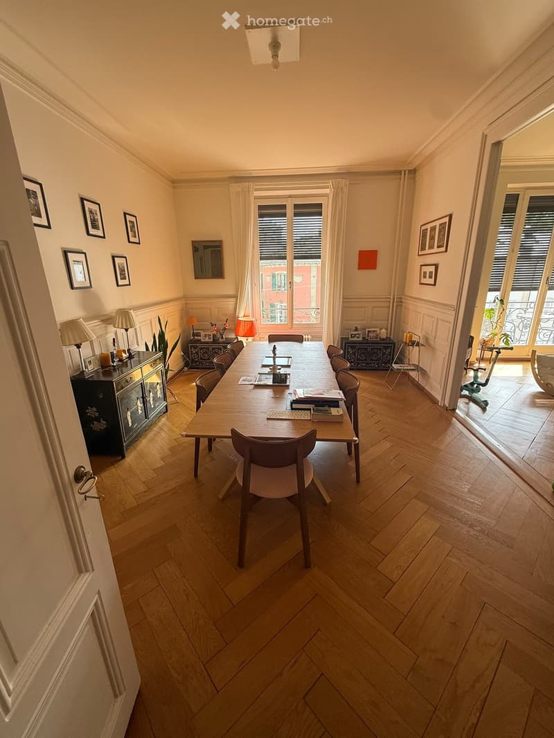 Spacious room with wooden herringbone flooring, large table surrounded by chairs, cabinets with lamps, framed pictures on walls, and a view of a balcony.