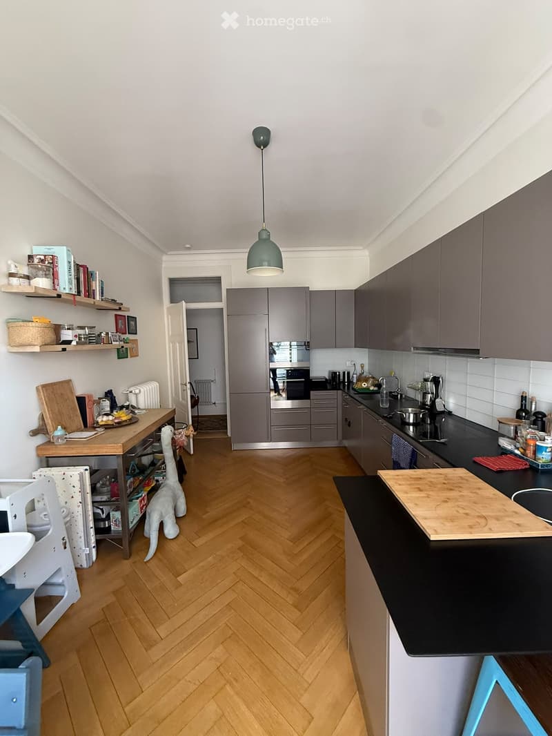 Modern kitchen with grey cabinets, stainless steel appliances, wooden herringbone flooring, and a central island with a black countertop. Shelves hold books and decorative items, and a small table with a chair is visible.