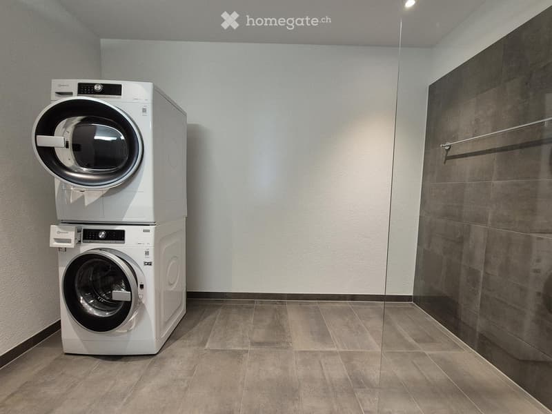 A small, modern laundry room with a stacked washer and dryer unit, tiled floor, and a glass shower enclosure.