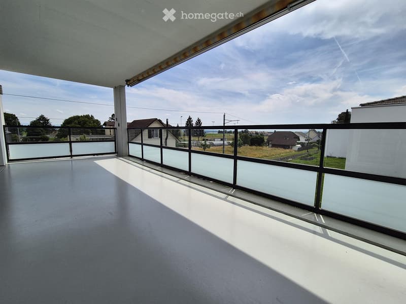 Empty balcony with modern glass railing, concrete floor, and scenic view of surrounding houses and fields.