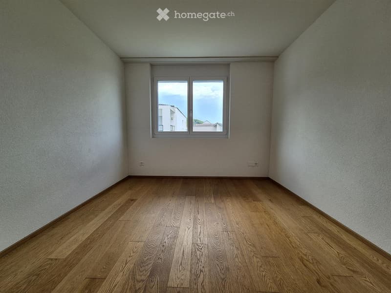 Empty room with wooden flooring and white walls, featuring a large window with a view of neighboring buildings.