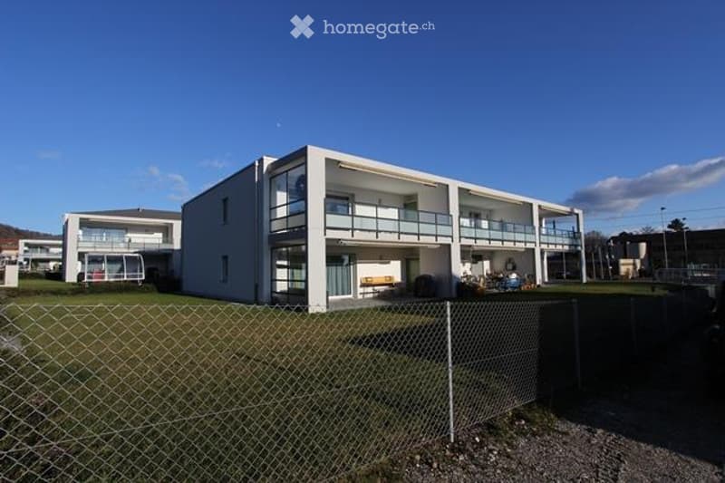 Modern two-story building with white exterior, multiple windows, and balconies. The building is surrounded by a fenced area with grass and gravel.