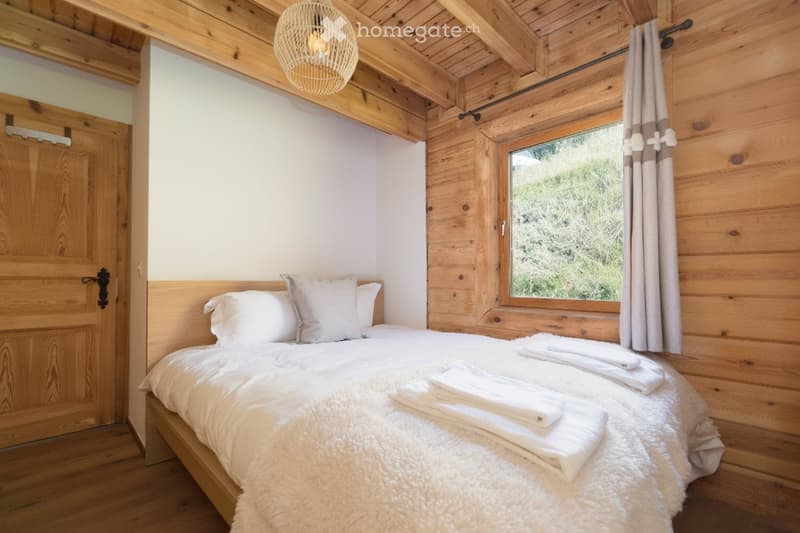 Cozy wooden bedroom with a large bed, white bedding, and a window with curtains. The room features exposed wooden beams on the ceiling and a hanging light fixture.