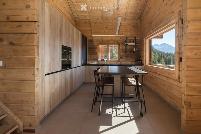 Modern kitchen with wooden walls and ceiling, large windows offering a mountain view, black countertop, and a small dining area with a table and stools.