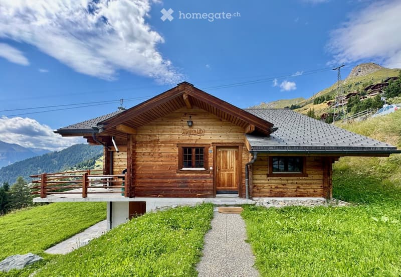 A wooden chalet with a sloped roof, stone foundation, and a small balcony overlooking a grassy area and mountain backdrop.