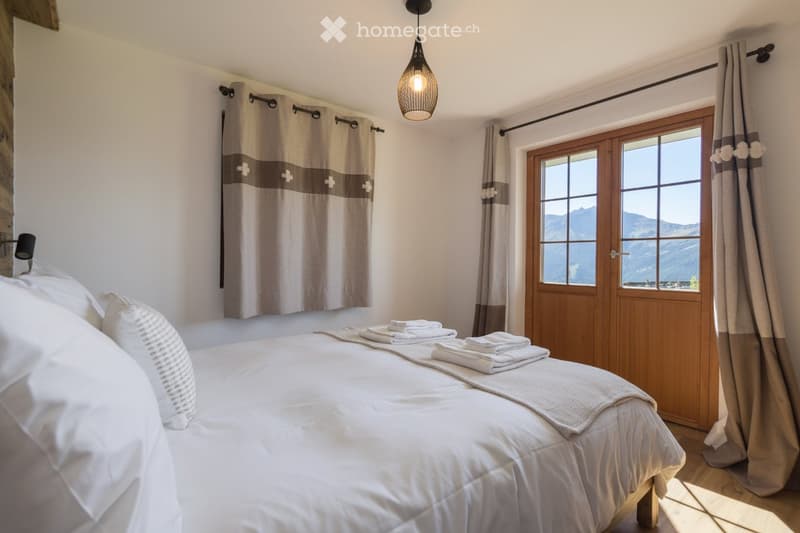 Cozy bedroom with a large bed, white linens, and a scenic mountain view through a wooden door and window. Features include a hanging pendant light and curtains.