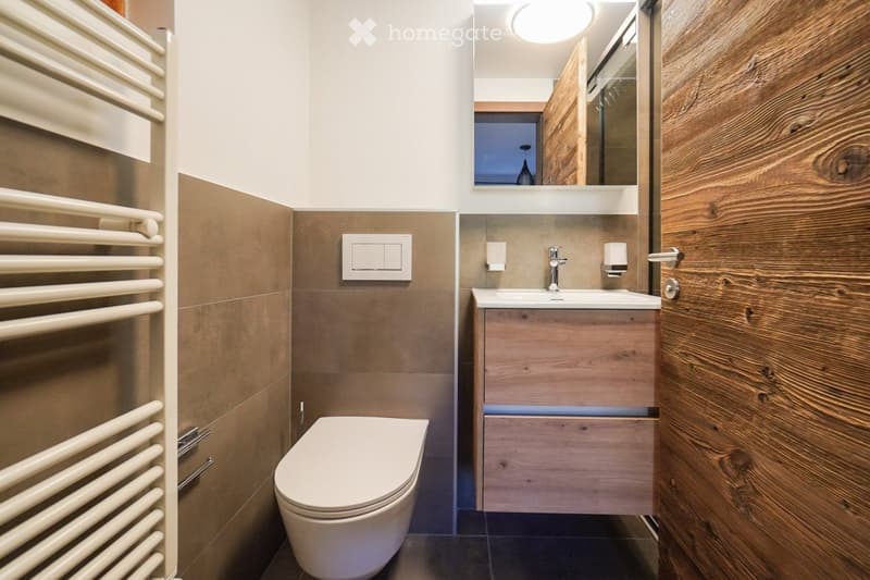 Modern bathroom with a toilet, sink, and mirror. The sink has a wooden cabinet underneath, and there is a radiator on the left wall.