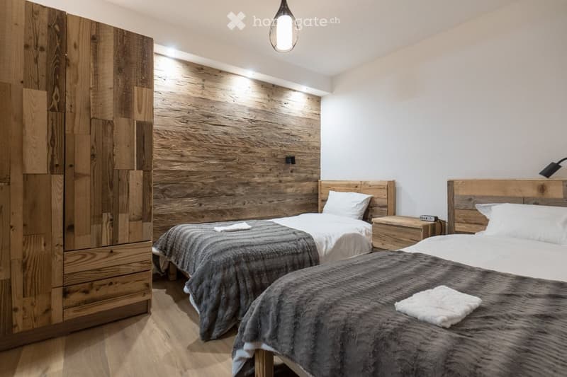 Cozy bedroom with two single beds, wooden nightstands, and a large wooden wardrobe. The beds are covered with grey fur throws and white pillows.