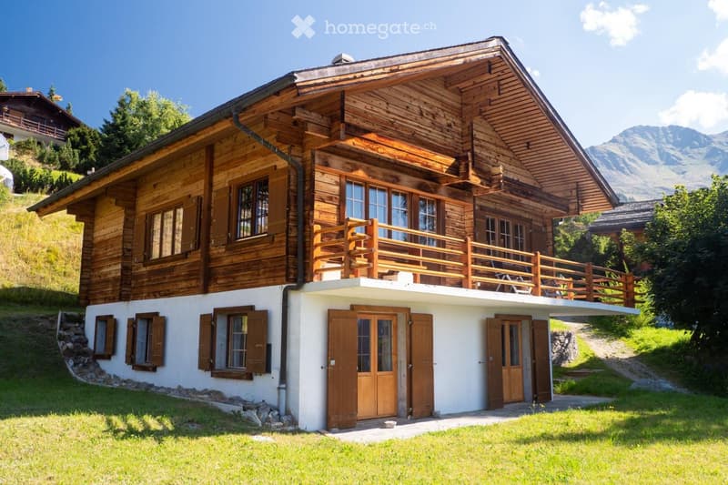 A two-story wooden house with a balcony, large windows, and a view of mountains in the background.