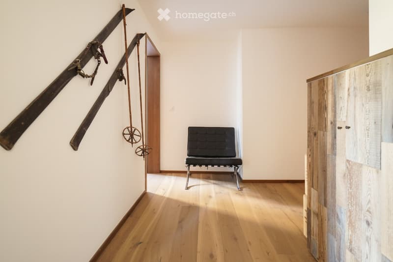 A narrow hallway with wooden flooring, white walls, and a black bench placed against the wall. A pair of skis and a decorative wheel are mounted on the left wall, and a wooden cabinet is visible on the right side.