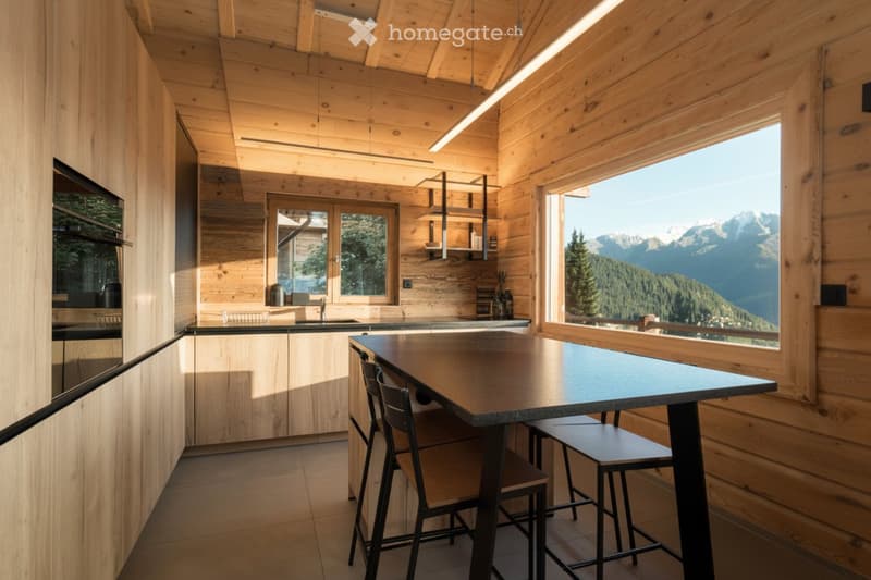 Modern kitchen with wooden walls, large windows offering mountain views, central island table with stools, and built-in shelves.