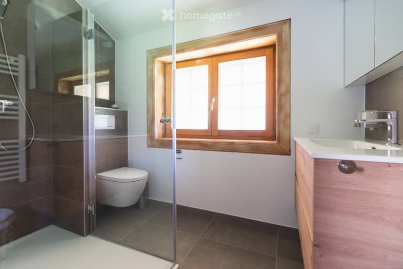 Modern bathroom with a glass-enclosed shower, toilet, sink with wooden cabinet, and a window with wooden frame.
