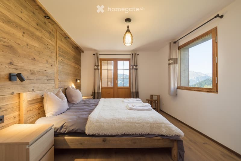 Cozy bedroom with wooden walls, a large bed with white pillows and a fluffy white throw, a nightstand, a chair, and large windows with curtains offering a mountain view.