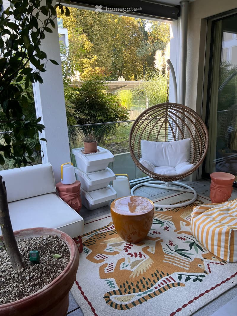 Balcony with a white sofa, wicker hanging chair, small table, colorful rug, and potted plants.