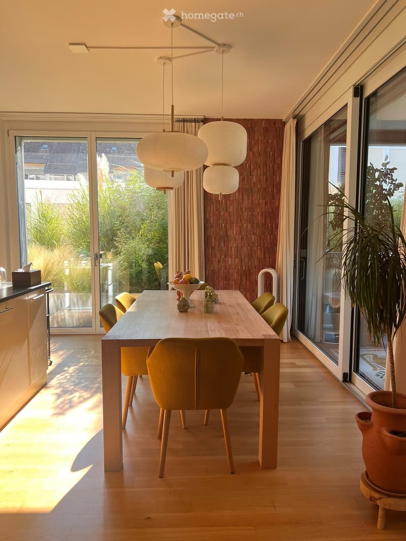 Bright dining area with a long wooden table, six green chairs, a fruit bowl, and a large potted plant near the window. The room features modern pendant lights and large glass doors leading to a garden.
