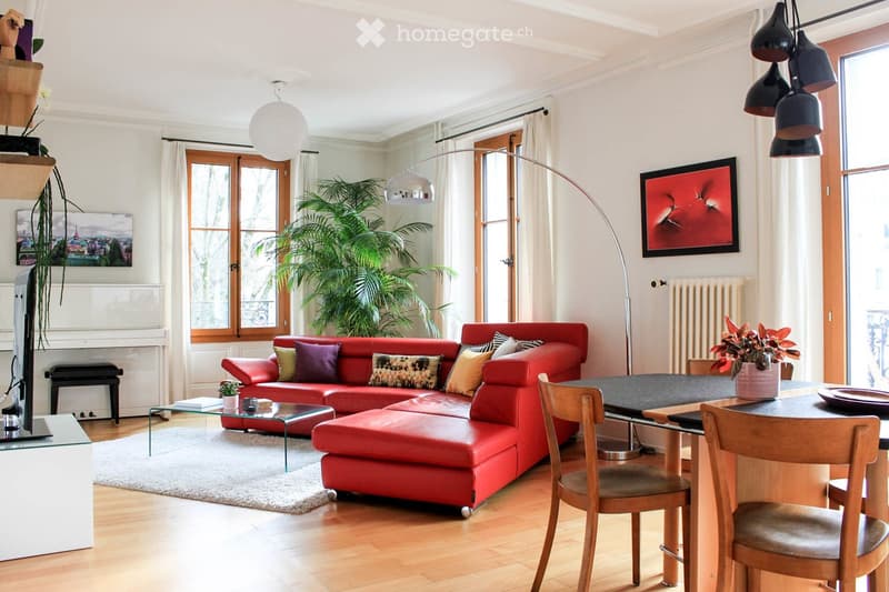 Modern living room with a red leather sofa, glass coffee table, wooden dining table with chairs, floor lamp, and large windows with curtains.