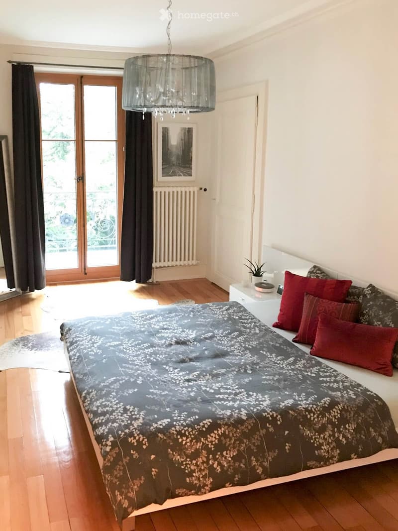 Bright bedroom with wooden flooring, large bed with grey and white patterned duvet and red pillows, nightstand with plant, chandelier, large window with dark curtains, radiator, and picture frame on wall.
