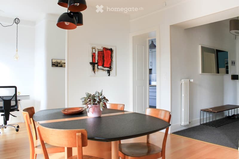 A modern dining area with a round black dining table surrounded by wooden chairs, a potted plant, and a bench. An office chair is visible to the left, and a mirror and radiator are on the right wall.