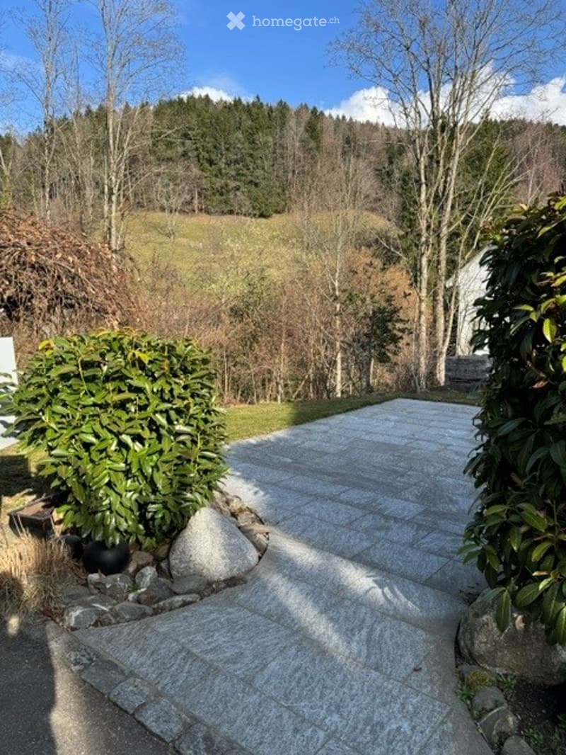 A paved outdoor area with a stone pathway leading to a house, surrounded by greenery and trees, with a scenic view of a forested hillside in the background.