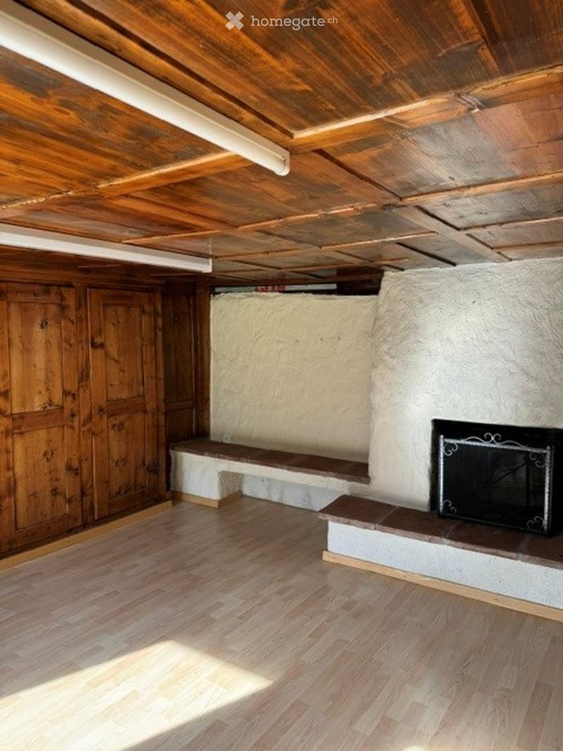 Empty room with wooden floor, exposed wooden ceiling, white plaster wall, and a fireplace with a black metal frame.