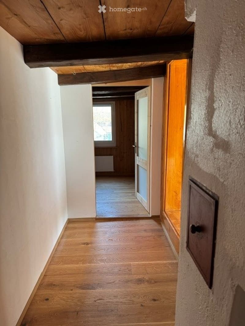 A narrow hallway with wooden flooring and exposed wooden beams on the ceiling. The walls are painted white, and there is a door at the end leading to another room.